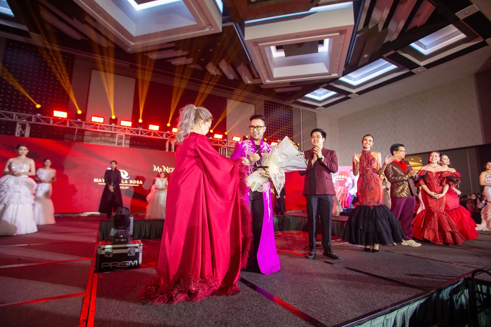 Guests on the gala stage under red and gold lights, applauding as a bouquet is presented