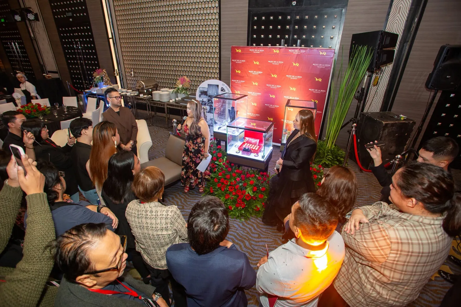 Guests gathered around glass display cases on a red-carpeted floor ringed with red roses, photographing the anniversary display
