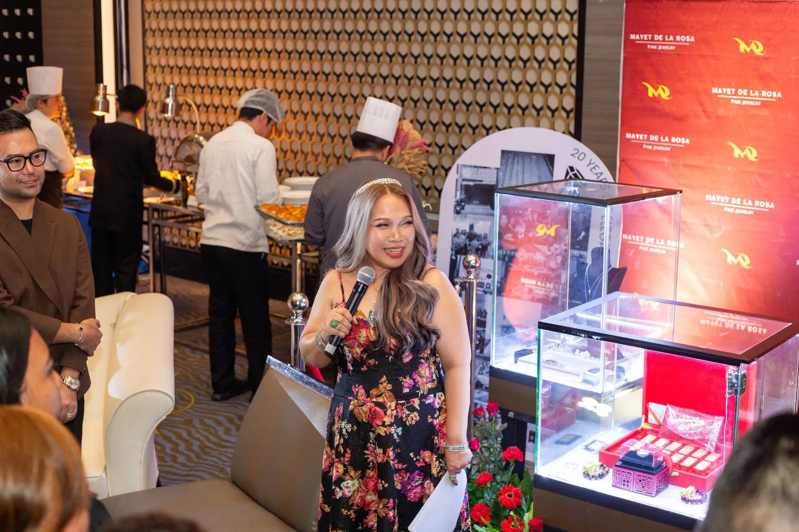 A speaker with a microphone addressing guests beside lit display cases of jewelry, in front of a red Mayet De La Rosa backdrop