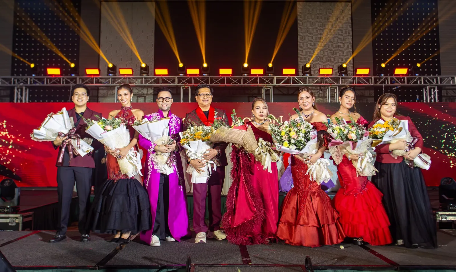Eight people holding bouquets on the gala stage, under gold stage lights against a red backdrop