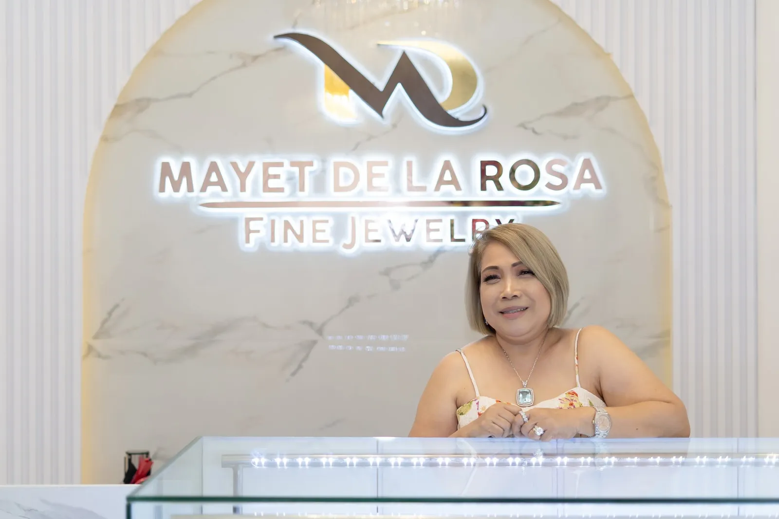 A smiling woman resting her hands on a glass jewelry counter beneath the Mayet De La Rosa Fine Jewelry sign